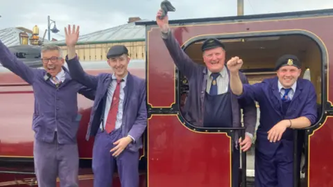 Four men, who are dressed in purple clothing, are standing in a row. Two are on the left and are standing outside of the locomotive, which is painted red and black with gold accents. Two men on the right are standing inside the locomotive. 
