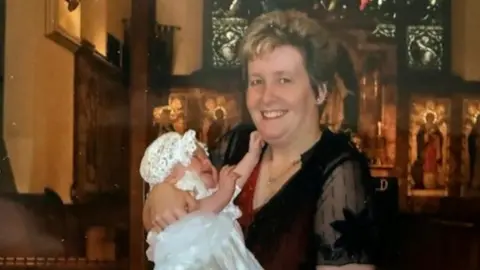 Family handout A scanned image of a middle-aged woman with short hair. She is standing in a church wearing a red dress with a mesh black top over the top. She is holding a crying baby wearing a white Christening dress and hat. 