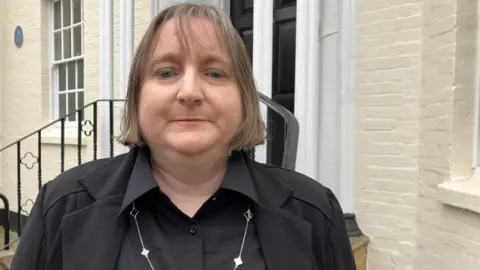 Grace Horton. She has short hair and is wearing black. She is looking at the camera and smiling. 