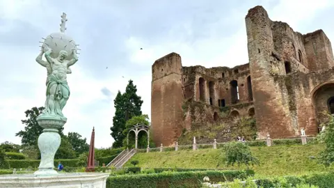 Kenilworth Castle, with several ruined red walls and a statue before it.
