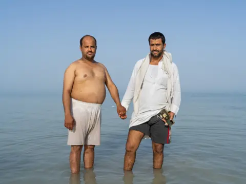 James Deavin Two men hold hands while standing in the sea