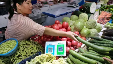 Getty Images A woman sells fruit and vegetables in a Chinese market stall with a QR code on display