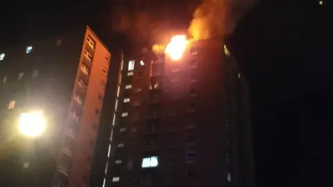 BBC Flames coming out of the16th floor of Twinnell House in Bristol after an e-bike started a fire. The building is seen on a dark night from street level, with some windows illuminated by internal lights