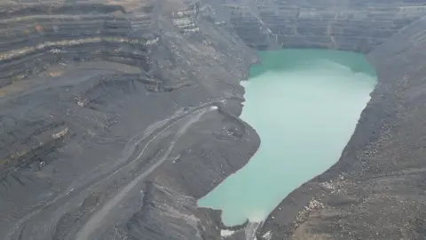 An aerial photograph of water pooling in the main void at Ffos-y-Fran mine. The water is bright blue, while grey cliffs walls surround it. 