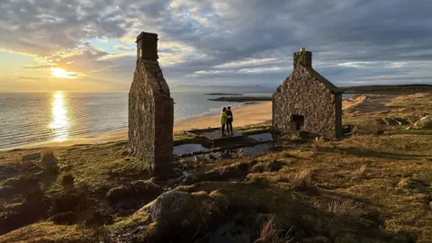 Richard Cooper Two people stand in between two gable ends of a house. The rest of the house is missing. There is a beach and sunset behind them.