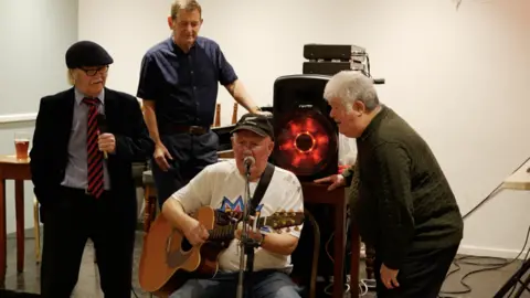 Provided Four people at the club performing a song around a microphone. One of them is playing the guitar.