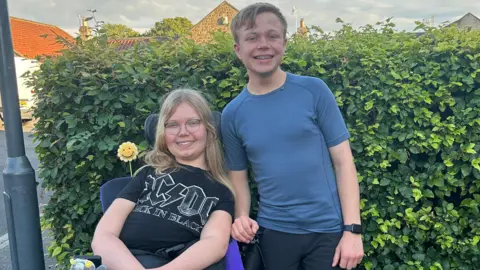 Two teenagers are next to each other. A girl, wearing glasses and an AC/DC T-shirt, is sitting in a wheelchair, with her brother, wearing a blue T-shirt, next to her. They are in front of a hedge on a residential street.