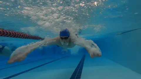 Swimmer with blue hat under the water with arms out and goggles on. Lanes are marked with dark blue lines