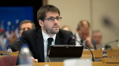 A head and shoulders picture of James Courtenay in a Southend City Council meeting, wearing a dark suit and tie, with a microphone and screen device in front of him