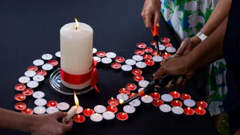 Getty Images Small red and white candles are arranged in the shape of an Aids ribbon. A large white candle is in the centre. The candles are being lit by some disembodied hands. 