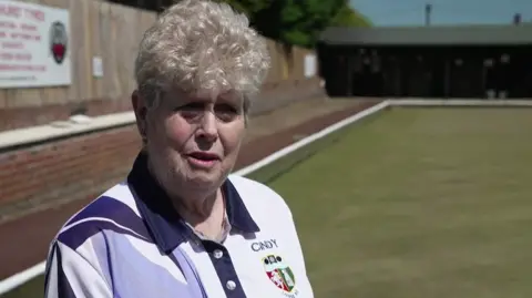 A woman with short curly white hair stands on a bowls lawn and wears a bowls kit t-shirt.