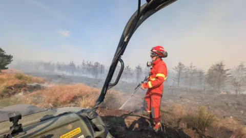 A firefighter in bright red protective clothing stands in a smoke-filled heathland, spraying water onto scorched ground after a fire. The scene is viewed from inside an off-road vehicle, with part of the vehicle frame visible in the foreground. Burnt vegetation and a line of smoke-shrouded trees stretch across the background under a pale blue sky.