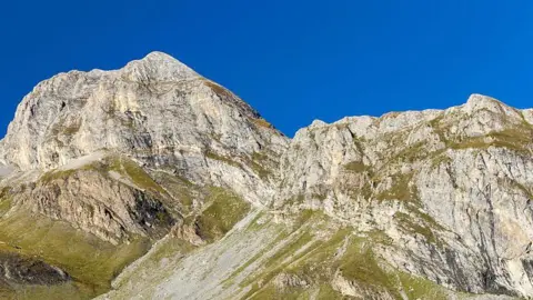 A large mountain with green vegetation at the bottom. The sky is bright blue behind it. 