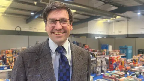 Fred Wyrley-Birch, a man with dark hair and spectacles, wearing a suit and tie, stands in front of a series of tables bearing boxed toys. He is smiling at the camera.