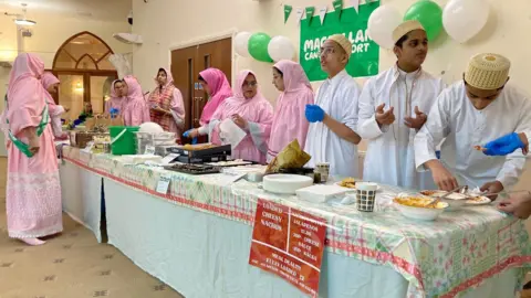 Men and women in traditional Dawoodi Bohra clothes running a charity cake stall.