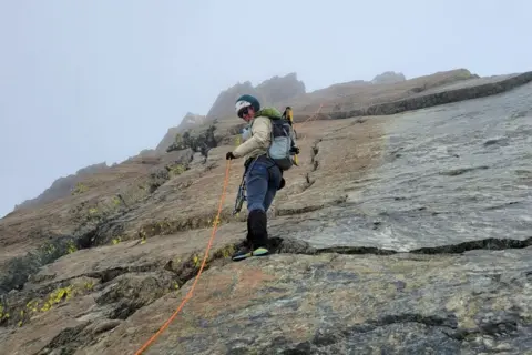 Rebekah Lee A woman in climbing gear with a helmet and rucksack climbs up a rock with a rope