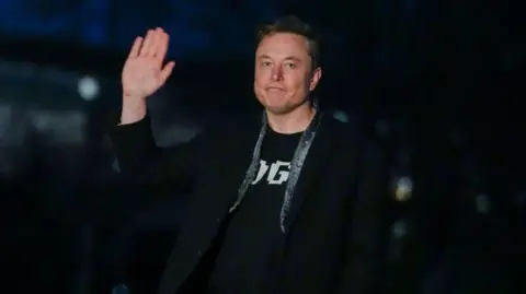 Getty Images Wearing a black jacket and black T-shirt, Elon Musk waves and smiles to the cameraas he walks across a lawn at night