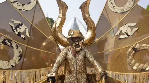 Emmanuel Adegboye / EPA A man wearing a gold sequin costume with a large manilla behind him holding up golden wings takes part in a festival in Calabar, Nigeria