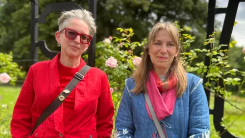 BBC/Aisha Iqbal Alison McIntyre, wearing a red top and jacket, stands next to Nicola Storr, who is wearing a blue jacket and pink scarf. They are standing outside in front of a metal gate surrounded by roses.