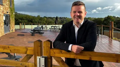 Anthony Hunt A man with brown hair, wearing a white shirt and dark jacket, leaning on a gate at a cafe.