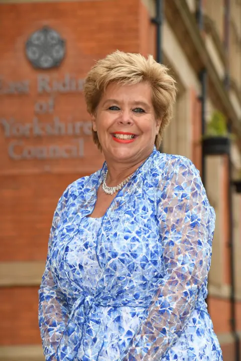 East Riding of Yorkshire Council A woman with short blonde hair stands in front of the ERYC building in Beverley, she wears bright lipstick, a string of peals, and a blue and white patterned blouse.