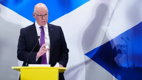 PA Media John Swinney standing at a yellow lectern with a saltire behind him.