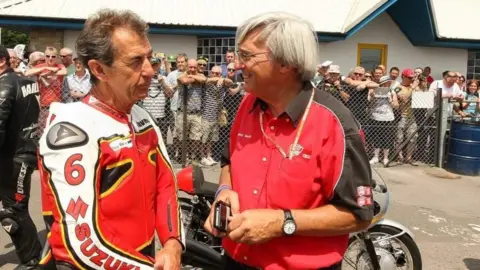 EDP Photo News A man wearing red leather Suzuki overalls standing next to Rodney Gooch, who is in a red and black shirt and smiling. There is a motorbike behind them and a crowd of people looking towards them from behind a barrier.