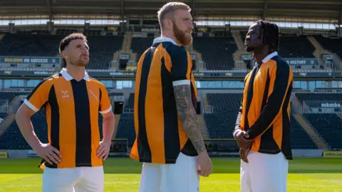 Three men stand on a football pitch wearing amber and black shirts with white shorts. They are all staring into the distance, looking in different directions. Behind them is a football stadium, with black seats in a stand.