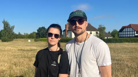 Holly Nichols/BBC Jess and Tim Roland standing in a field with two detached houses behind them. Jess is wearing dark glasses and a black T-shirt and Tim is wearing a green baseball cap, dark glasses and a white T-shirt.
