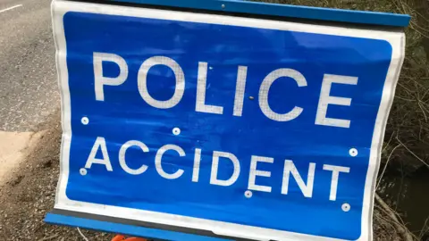 A blue "Police accident" sign on the verge of a road.
