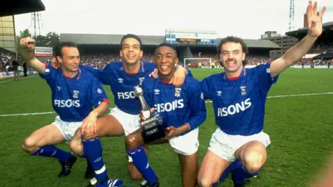 Ben Radford/Getty Simon Milton, Jason Dozzell, Chris Kiwomya and John Wark crouching on the pitch at Portman Road in their Ipswich Town kits, with the old Second Division trophy being held by Kiwomya.