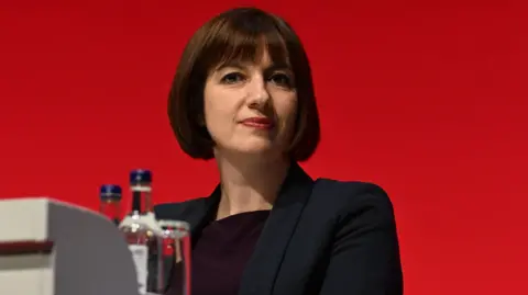 Education secretary Bridget Phillipson, who has short brown hair and a fringe, wears a black blazer and dark top and is pictured against a red background. Bottles and a glass of water are seen in the foreground.
