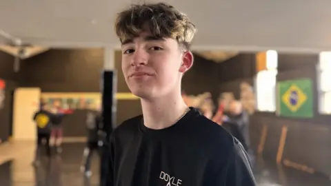 A boy with brown hair is turned sideways smiling into the camera, he is dressed in a black t-shirt. Behind him is an out-of-focus martial arts studio.