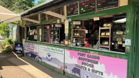 A small cafe with three small hatch openings. The structure is mostly green with black boards pinned up above the openings with prices and items written on them. 