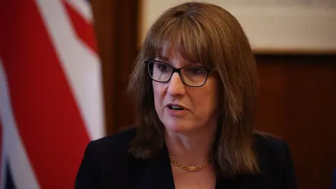 Head and shoulders shot of Rachel Reeves wearing a black jacket, gold necklace, and black glasses
