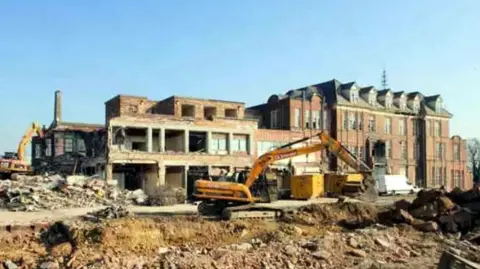United Lincolnshire Hospitals NHS Trust A yellow bulldozer digs through rubble in front of part-demolished, red-brick hospital buildings at Lincoln County Hospital.