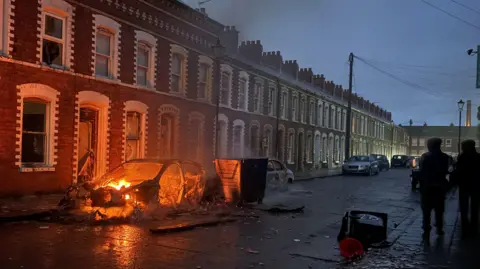 Reuters A burned-out car sits outside a row of terraced houses, with smoke still rising from the wreckage. Debris is scattered across the street as a small number of people stand nearby.