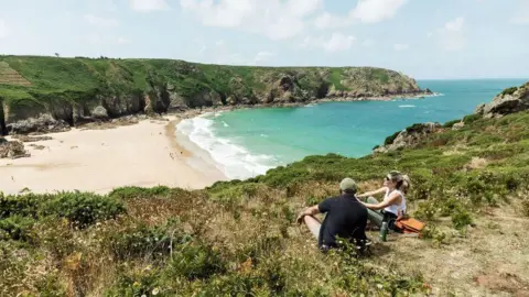 Visit Jersey A man and a woman are sitting near a cliff edge leading to a white sandy beach below and a clear green-coloured sea on a sunny day. On the opposite side of the small bay there is a grassy headland. There are a couple of people on the beach.