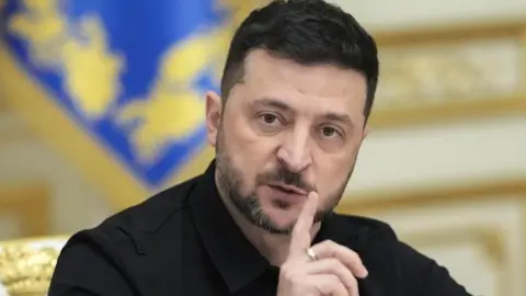 A head and shoulders image of Volodymyr Zelensky wearing his usual black suit and holding his index finger near his mouth. He has a near  beard and moustache.