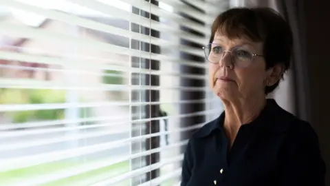 Sean De Francesco/Firecrest Films Carol Gillies looking out of a window. She has short hair and glasses and is wearing a black top.