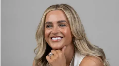 Shows a woman with blond hair smiling. She is wearing a wedding ring and a cream top