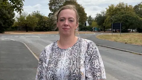 Kirstie stands on a pavement with a junction behind her, looking at the camera. She wears a grey, patterned top