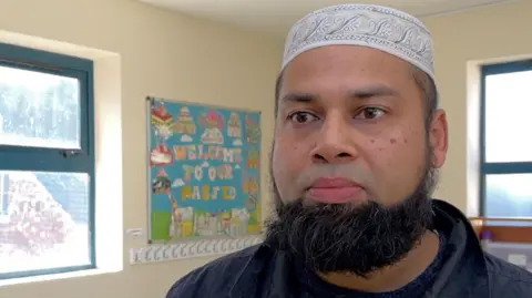 Bulbul Miah stands inside the Jame Masjid mosque. He is wearing a skullcap and has a black beard. In the background, a colourful sign reads 