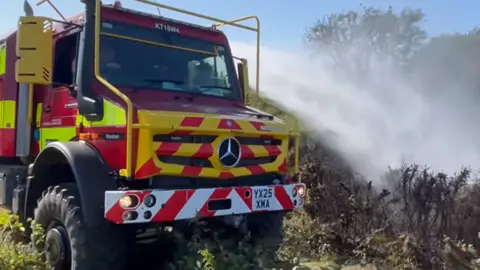 DWFRS A large fire fighting vehicle driving through gorse spraying water from jets on its far side