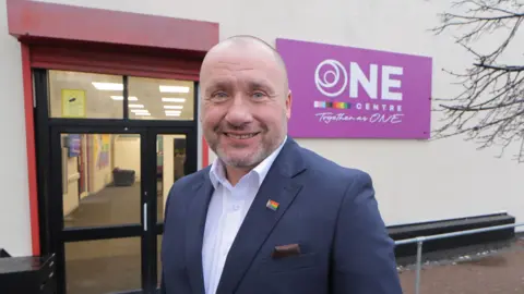 Out North East Peter Darrant, CEO of charity OUT North East stands in from of the ONE Centre in Pelaw Gateshead. He is smiling, wearing a suit jacket with a Pride flag pin on the lapel, and white shirt.