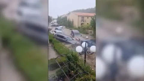 Cars being washed away in brown flood water, through residential buildings