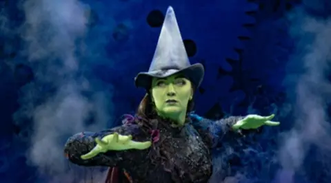 An actor is playing Elphaba in Wicked, with a black witch's hat and dress and her skin is painted green. She has her arms slightly raised as if casting a spell. There is stage smoke in the background.