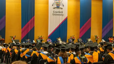 Cranfield University An indoor graduation ceremony at Cranfield, which has a hall full of students wearing black mortar board hats and black robes with a blue and orange hood. On the stage there are floral displays and academics in their university robes and hats.