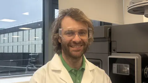 Manchester Metropolitan University Dr David Megson smiles at the camera. He is in a lab and there is a metal machine behind him. He has long shoulder-length curly brown hair and facial hair. He is wearing a green polo top and a white lab coat and protective glasses. 