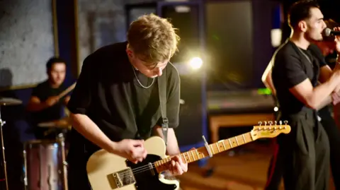 Jamie Niblock/ BBC Guitarist Luke Ellis, mid strumming his electric guitar which is two tone white and black.